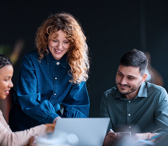 A woman stands smiling, showing her laptop screen to seated coworkers